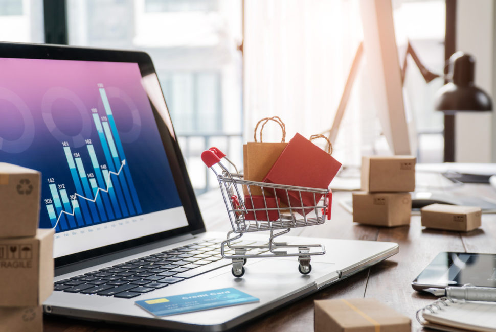 Shopping bags in shopping cart and credit card on laptop with paper boxes on table and sales data economic growth graph on screen, buying and selling services online on network, online shopping and e-commerce concept